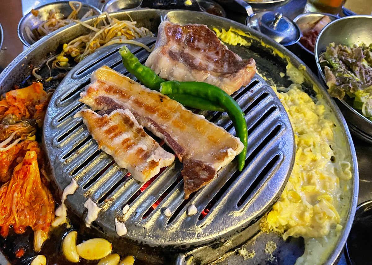 Gebratenes Kimchi in einer Pfanne neben Samgyeopsal auf dem Korean BBQ Grill
